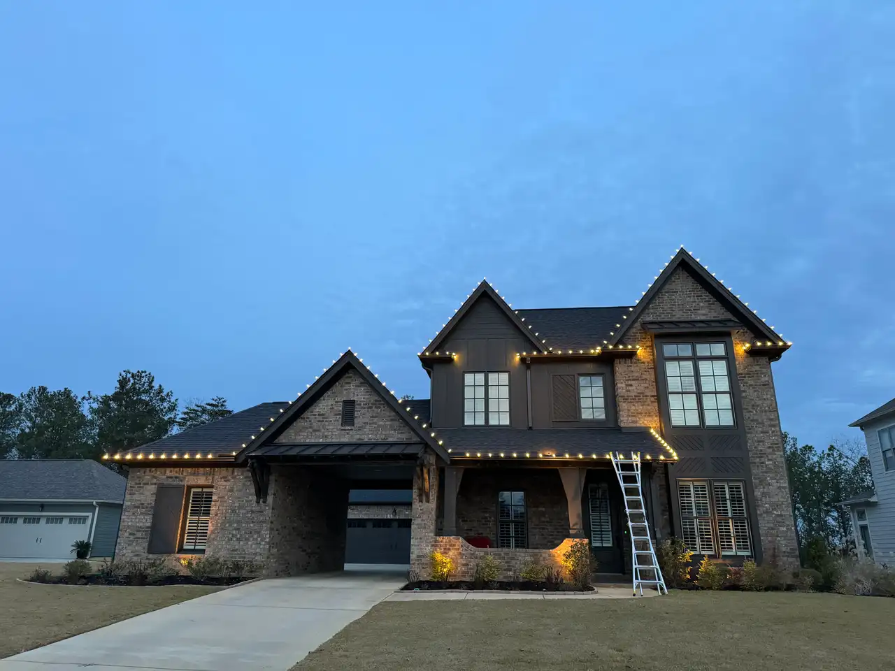 Two-story brick home at dusk with warm white lights outlining the roof peaks and gables, with a ladder at the front porch